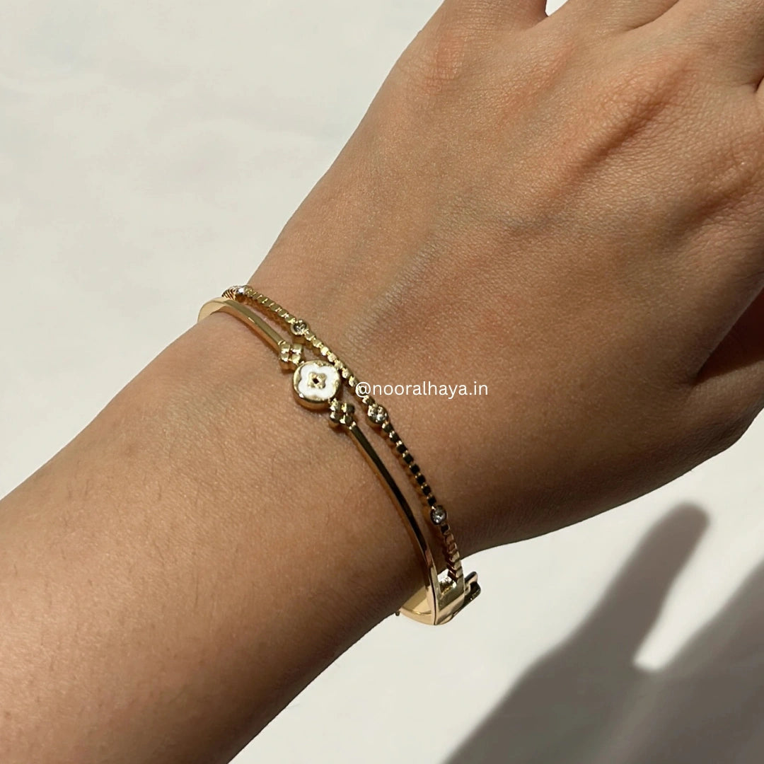 A close-up of a person's wrist wearing two delicate gold bracelets, one with a small central floral charm and the other with a textured chain, on a light neutral background.