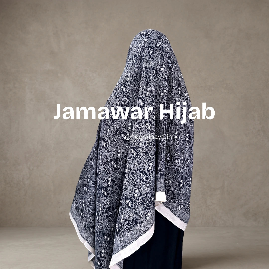 A woman wearing a Jamawar Hijab, shown from the back. The hijab is a dark gray with an intricate white paisley and floral pattern, finished with a white border by noor al haya