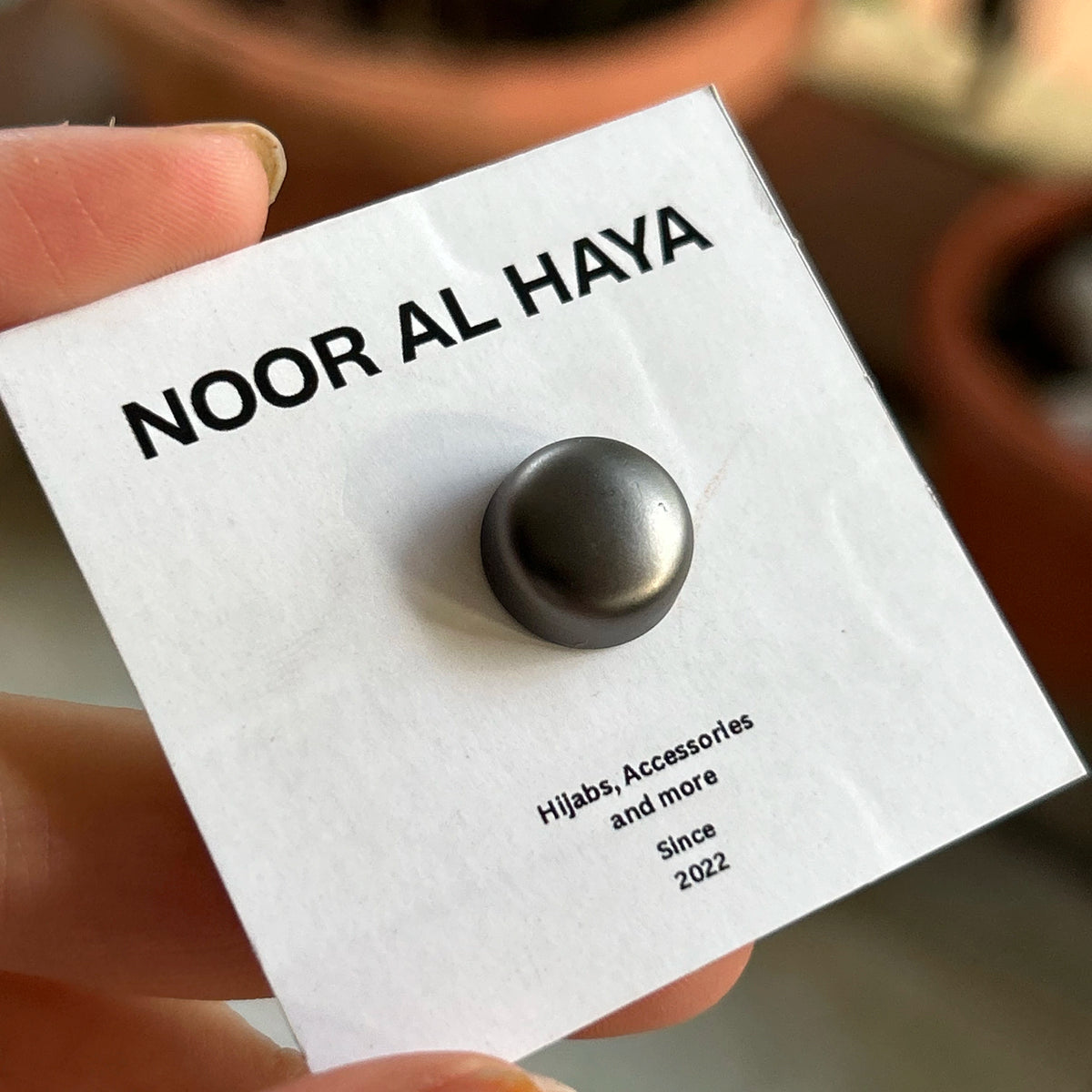 A close-up of a hand holding a white display card featuring a single round dark metallic button or stud. The card is branded 'NOOR AL HAYA' and includes the text 'Hijabs, Accessories and more Since 2022'.