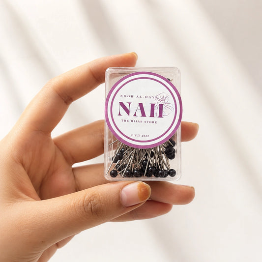 A hand holding a small clear plastic box filled with black-headed sewing pins. The box has a round purple and white label that reads 'NAH THE HIJAB STORE'.