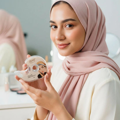 A woman wearing a dusty pink hijab holds a roll of ZERA Hijab Styling Tape in front of her. The brand name 'ZERA' is visible in the top left corner, and a mirror in the background shows her profile.