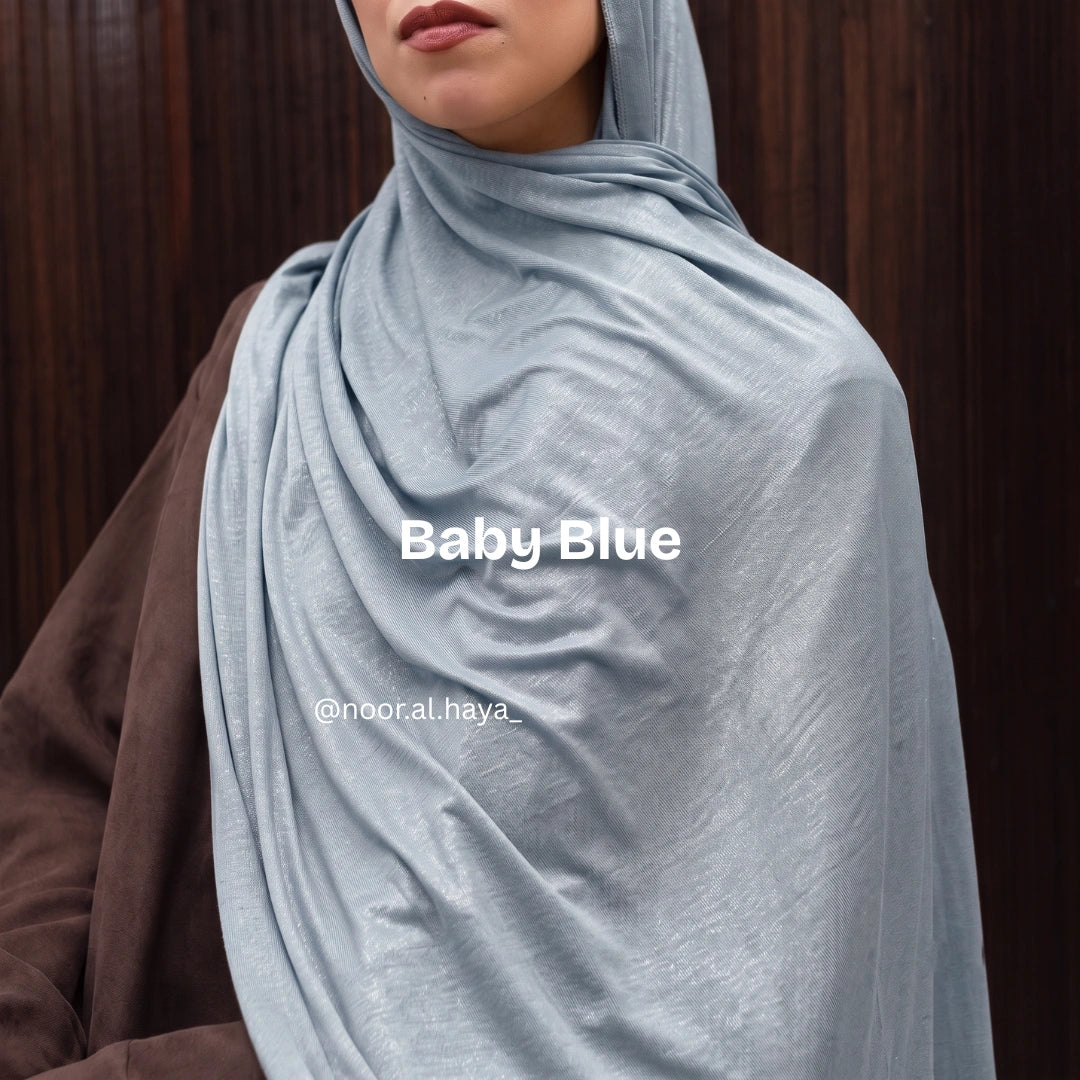 A close-up of a person wearing a soft baby blue hijab or shawl draped around the neck and shoulders, with a brown garment visible on the left side. The image includes the text 'Baby Blue' by noor al haya