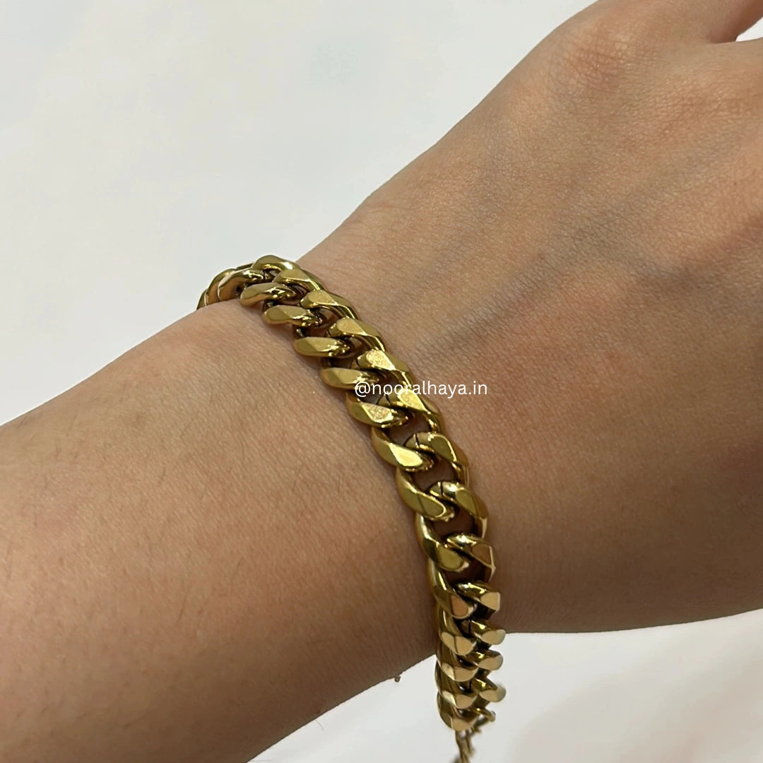 A close-up of a gold-toned Cuban link chain bracelet worn on a mode's wrist against a plain light background by noor al haya