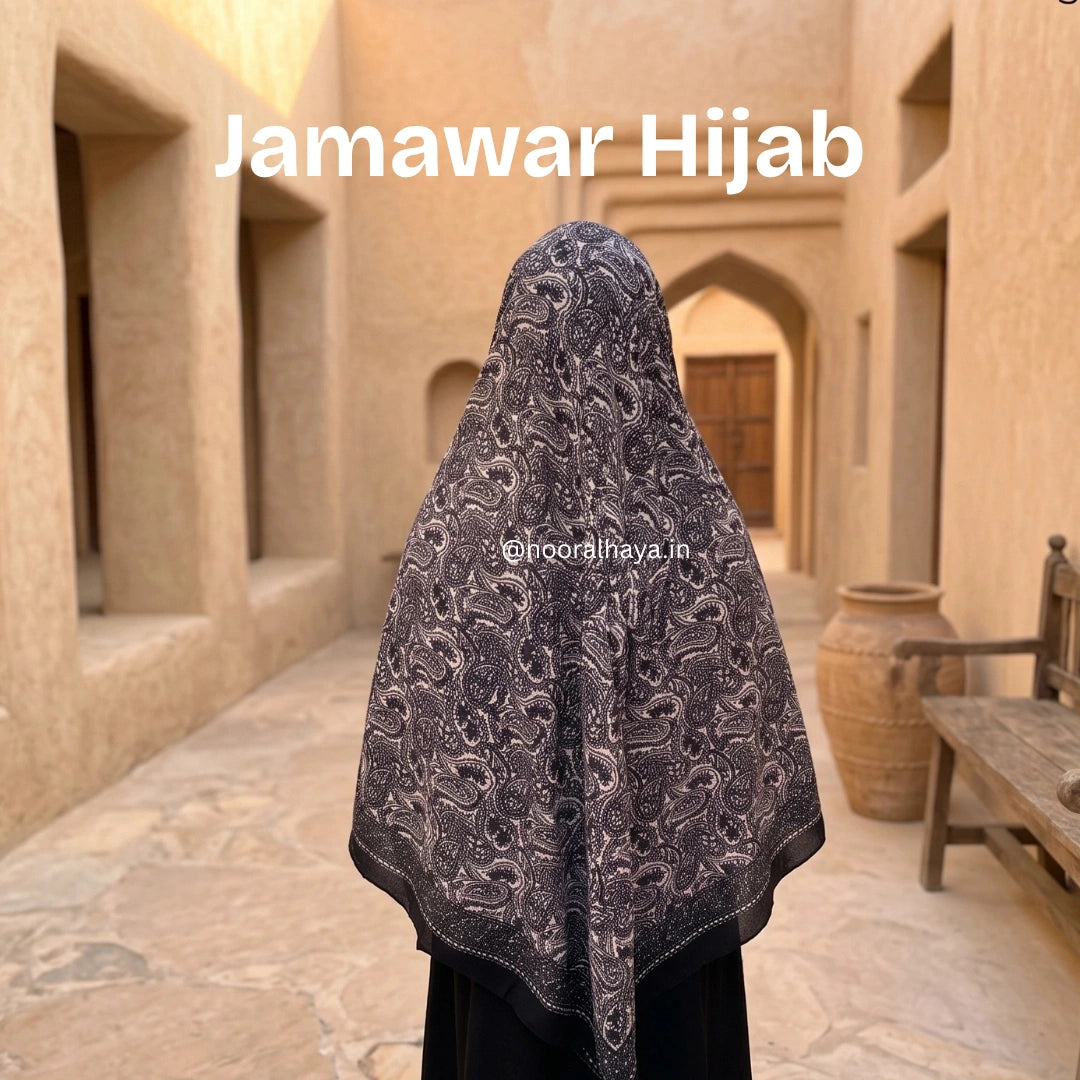 A woman viewed from behind wearing a dark gray and black paisley-patterned hijab in a traditional courtyard. The text 'Jamawar Hijab' is displayed at the top, and a watermark 'nooralhaya.in