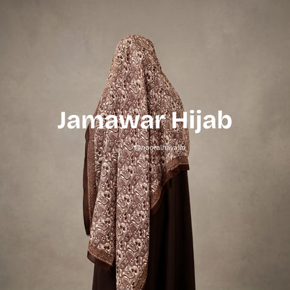 A woman wearing a brown embroidered hijab with floral patterns, shown from the back against a textured gray background. White text overlay reads 'Jamawar Hijab' with a smaller watermark 'jamawar hijab' below it. a model wearing jamawar hijab by noor al haya