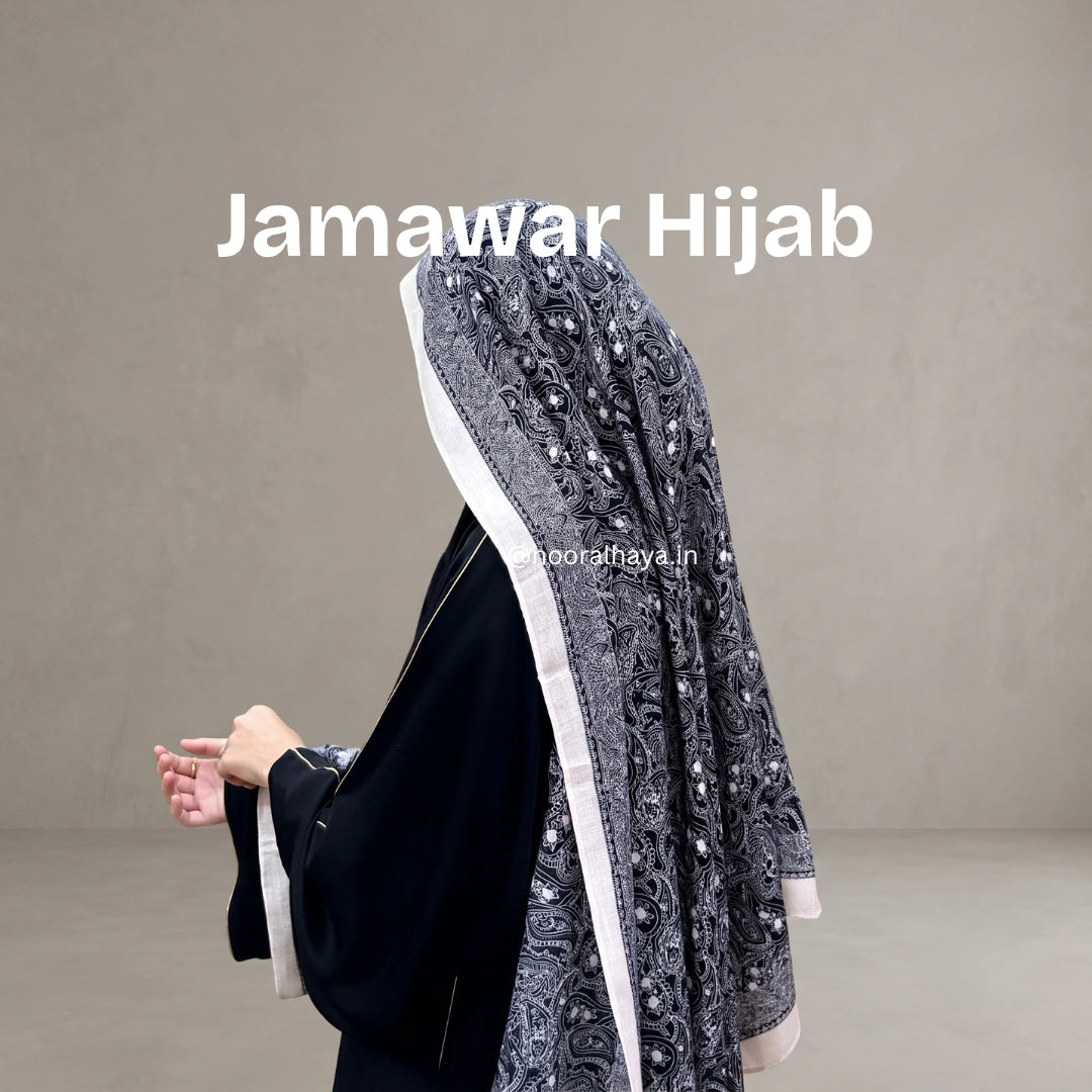 A woman wearing a black abaya and a dark gray patterned hijab with white borders, shown from the back against a plain gray studio background. The text 'Jamawar Hijab' is overlaid at the top by noor al haya