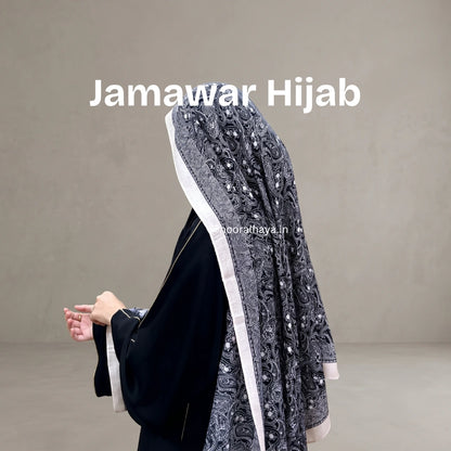 A woman wearing a black abaya and a dark gray patterned hijab with white borders, shown from the back against a plain gray studio background. The text 'Jamawar Hijab' is overlaid at the top by noor al haya