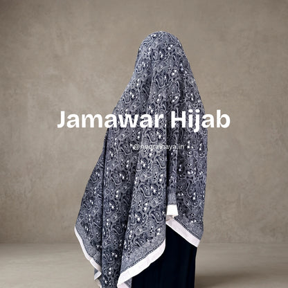 A woman wearing a Jamawar Hijab, shown from the back. The hijab is a dark gray with an intricate white paisley and floral pattern, finished with a white border by noor al haya