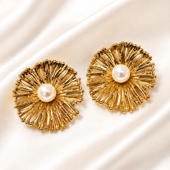 A pair of gold-toned statement earrings with a textured, sunburst radial design and a central white pearl, displayed on a draped white satin fabric.
