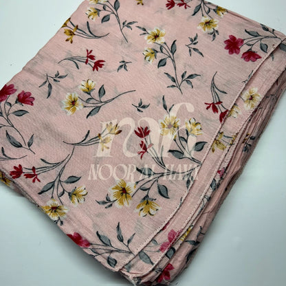 A folded light pink fabric item, likely a scarf or dupatta, featuring a delicate floral pattern with yellow, red, and white blossoms and gray-green leaves. A semi-transparent watermark reading 'NOOR AL HAYA' is overlaid across the center by Noor Al Haya